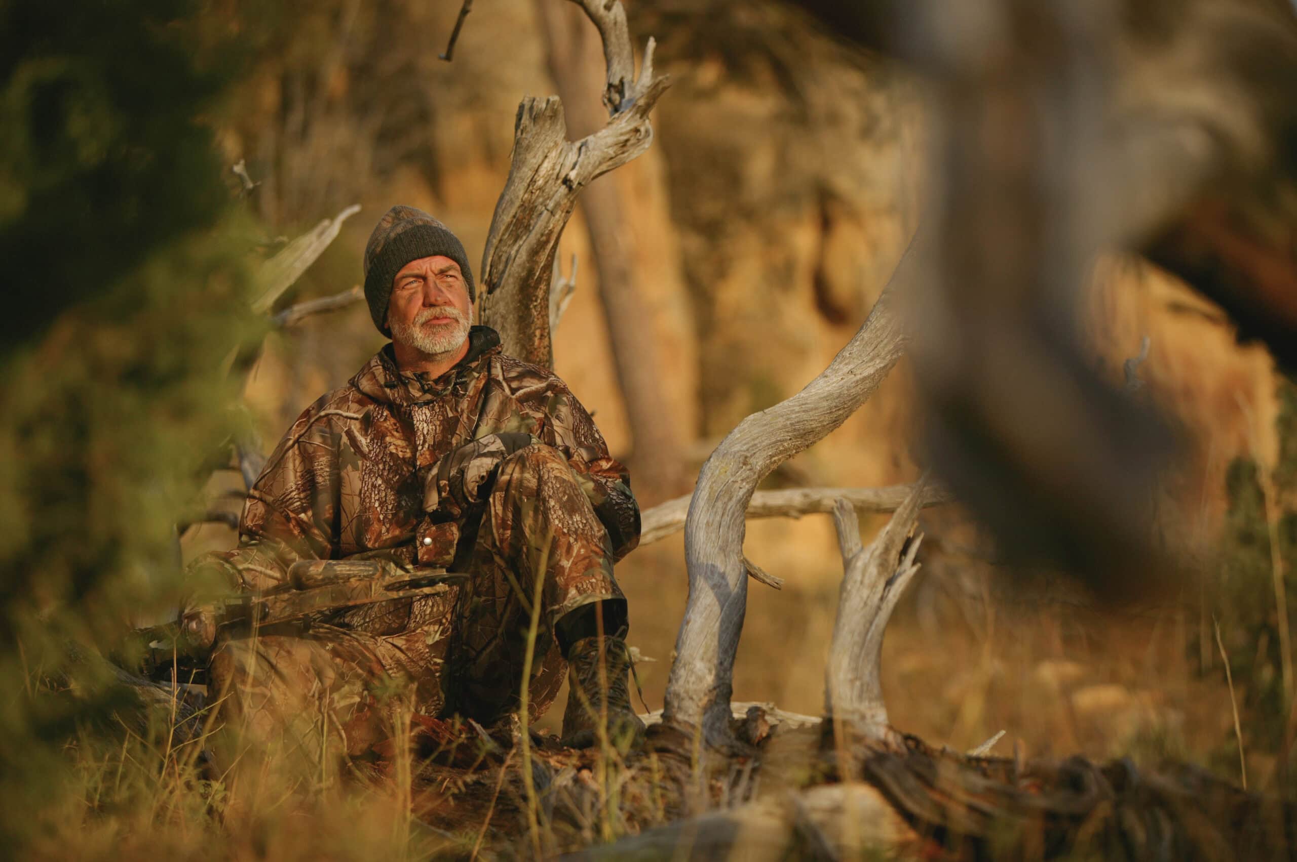 Man in camouflage gear sitting quietly in a wooded area during a turkey hunting trip, surrounded by dry grass and tree limbs at golden hour.