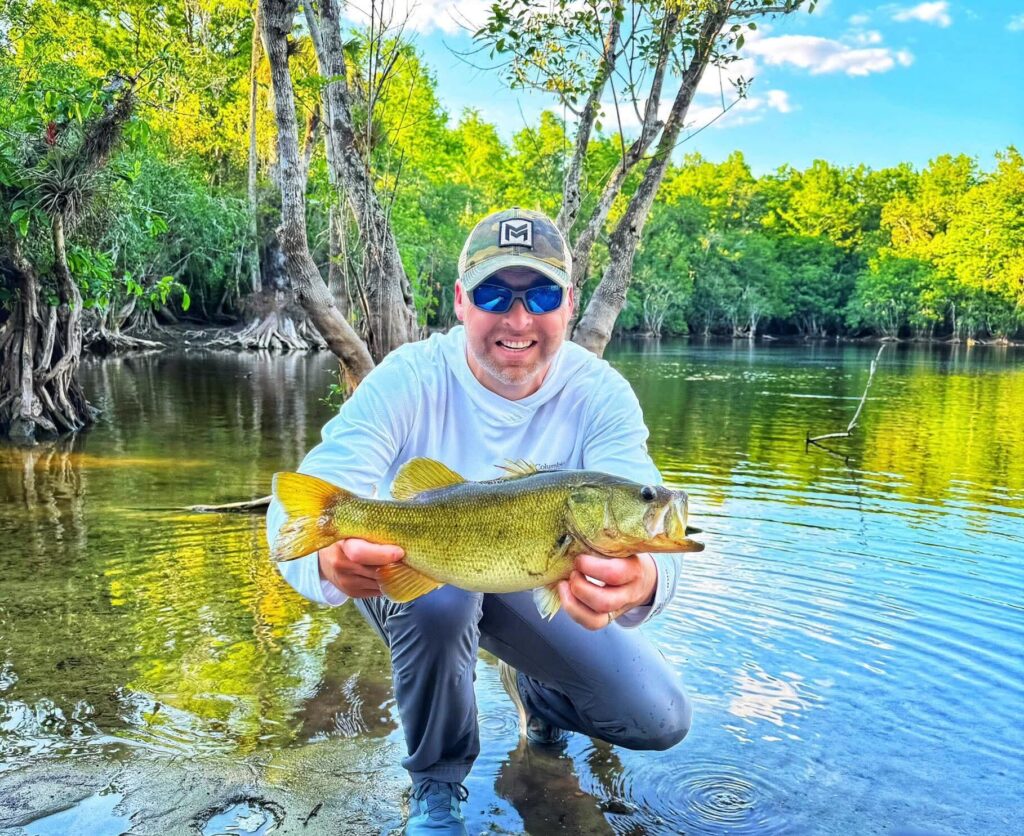 John Douglas holding a largemouth bass on the riverbank, a great example of what is fly fishing in a scenic, wooded environment.
